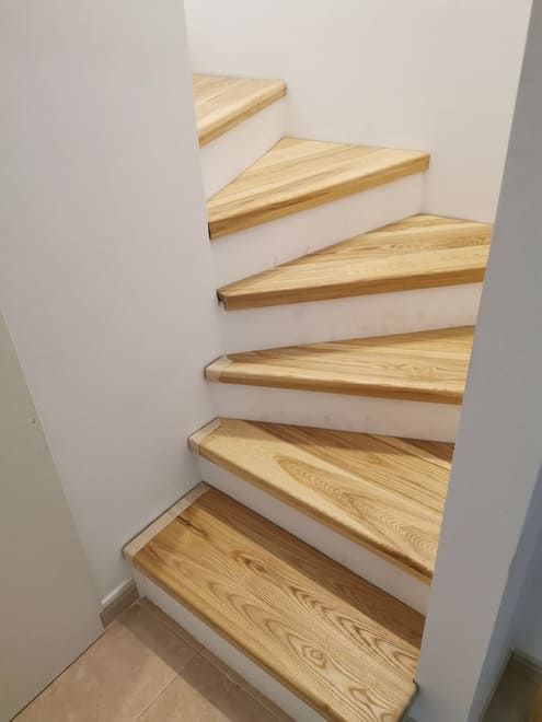 Escalera curva de madera con peldaños de madera clara y paredes blancas en un hueco de escalera estrecho.