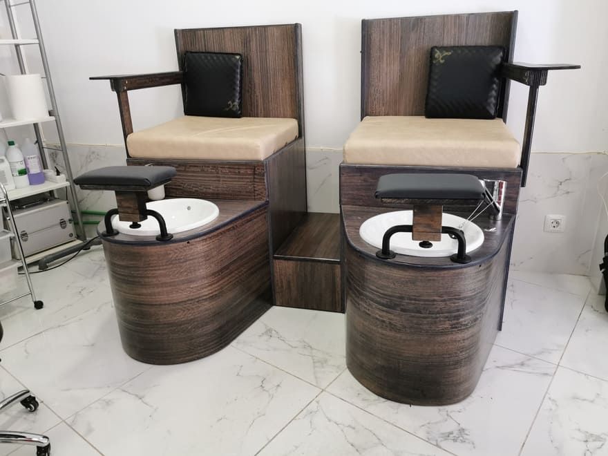 Dos sillones de pedicura modernos con bases de madera oscura en una sala de salón, cada uno con lavabo para los pies y reposabrazos.
