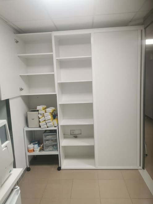 Armario blanco abierto con estantes y un cajón en una habitación con suelo de baldosas, que contiene diversos artículos de primera necesidad.