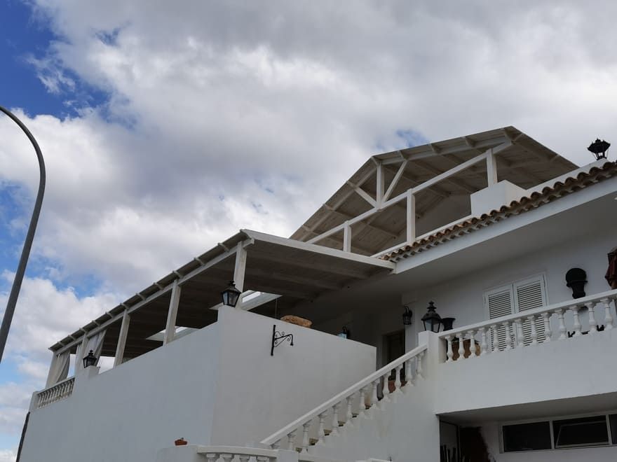Edificio de estuco blanco con techo de tejas rojas y terraza abierta en la azotea contra un cielo nublado.