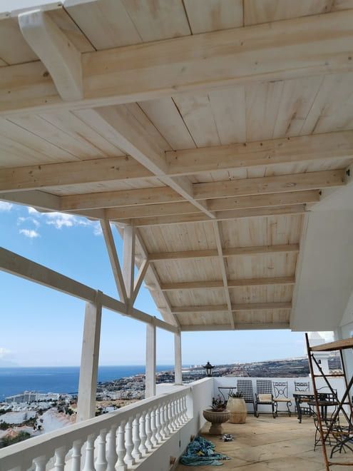 Terraza cubierta junto al mar con vigas de madera, barandilla blanca y sillas, con vistas al océano y al pueblo.
