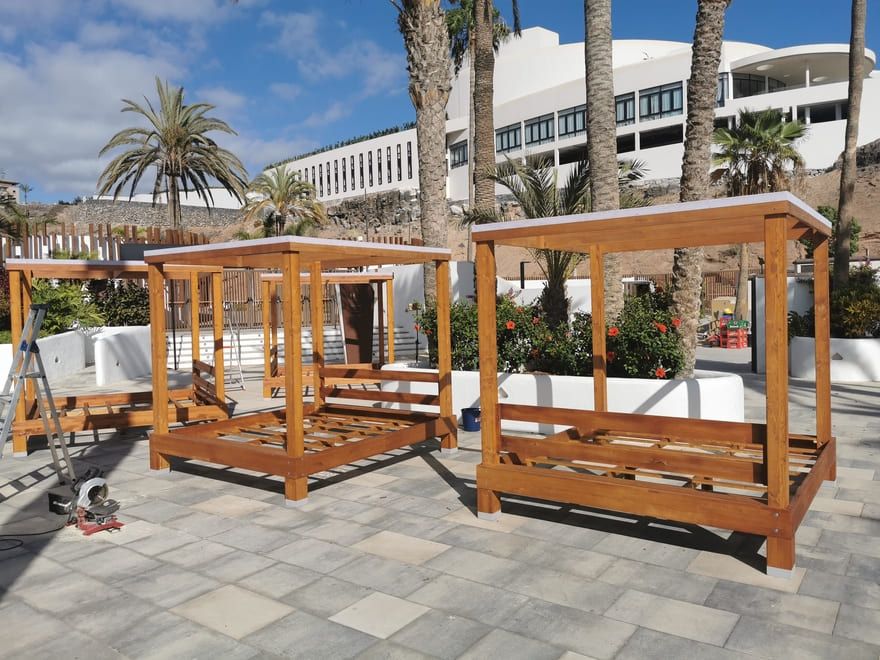 Tres pérgolas de madera con asientos en una plaza exterior soleada con palmeras y un edificio del complejo turístico.