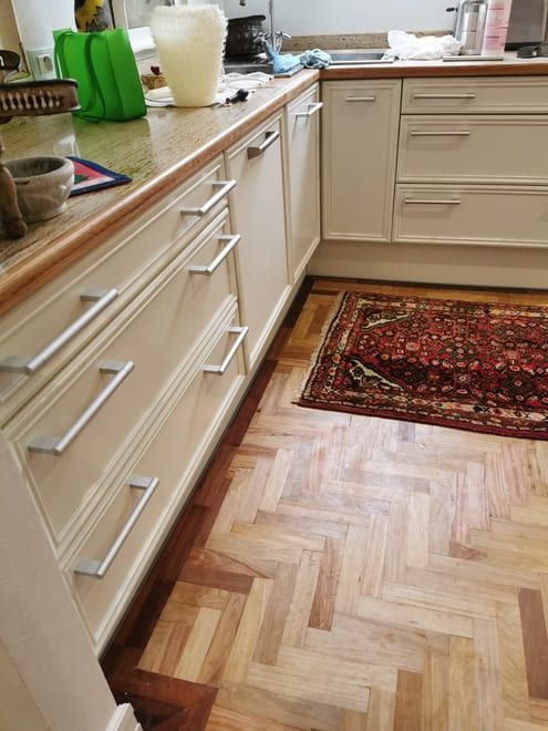 Cocina con armarios de madera clara, encimeras y una pequeña alfombra roja estampada sobre suelo de madera.