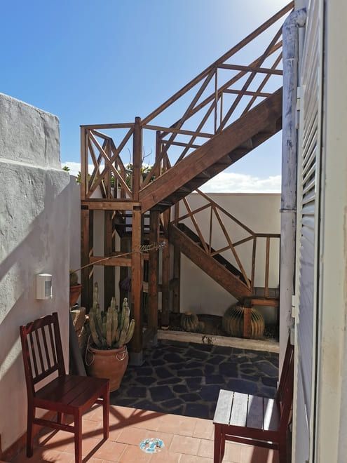 Pequeño patio soleado con escaleras de madera, paredes blancas, plantas en macetas y una silla sobre un suelo de piedra.