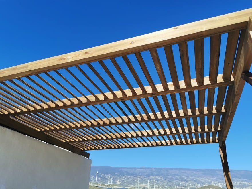 Techo de pérgola de madera con vigas de listones contra un cielo azul claro, con vistas a una ladera lejana.