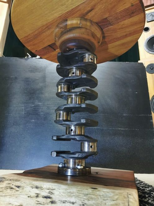 Primer plano de un cigüeñal metálico con un disco de madera arriba y otro abajo, montados sobre un fondo oscuro.
