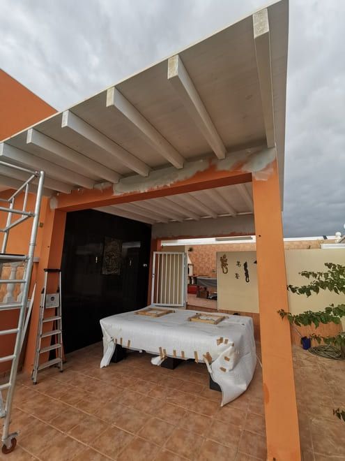 Patio con pérgola cubierta, escalera y mesa bajo un techo con detalles naranjas en un jardín trasero.