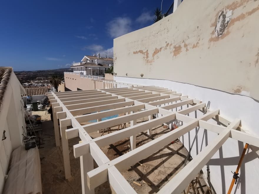 Terraza en construcción en la azotea con vigas de madera blanca, paredes beige y cielo azul con vistas a la ciudad.