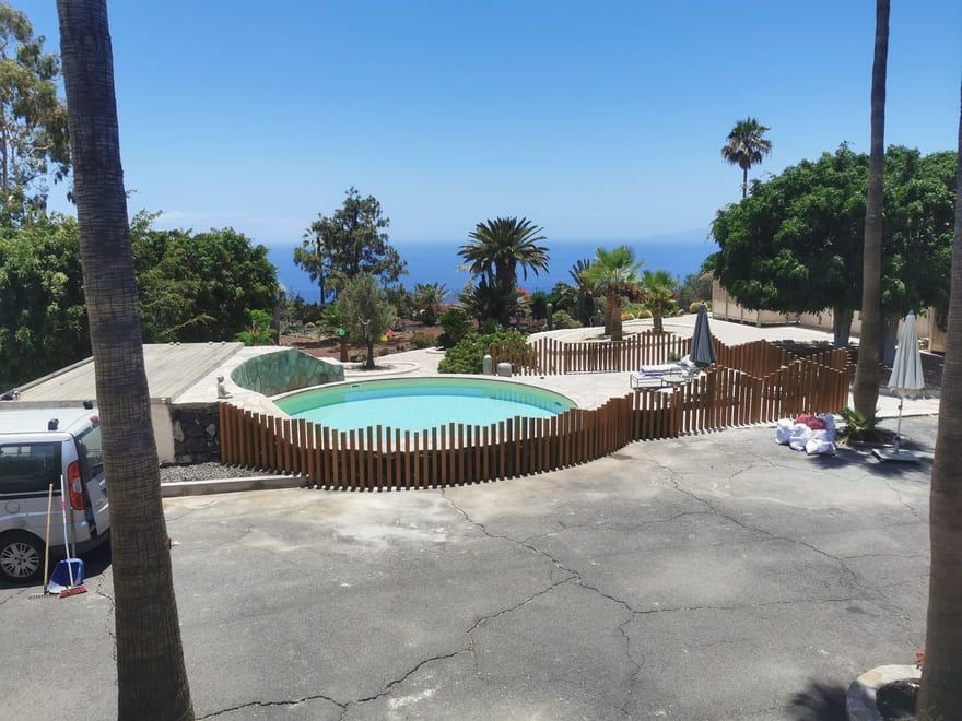 Piscina vallada con vistas al mar, palmeras y un coche aparcado en una ladera soleada.