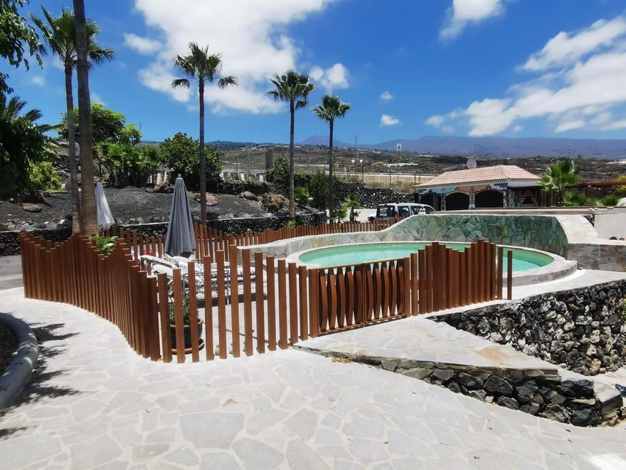 Piscina del complejo con jacuzzi vallado, palmeras y cielo azul junto al océano.