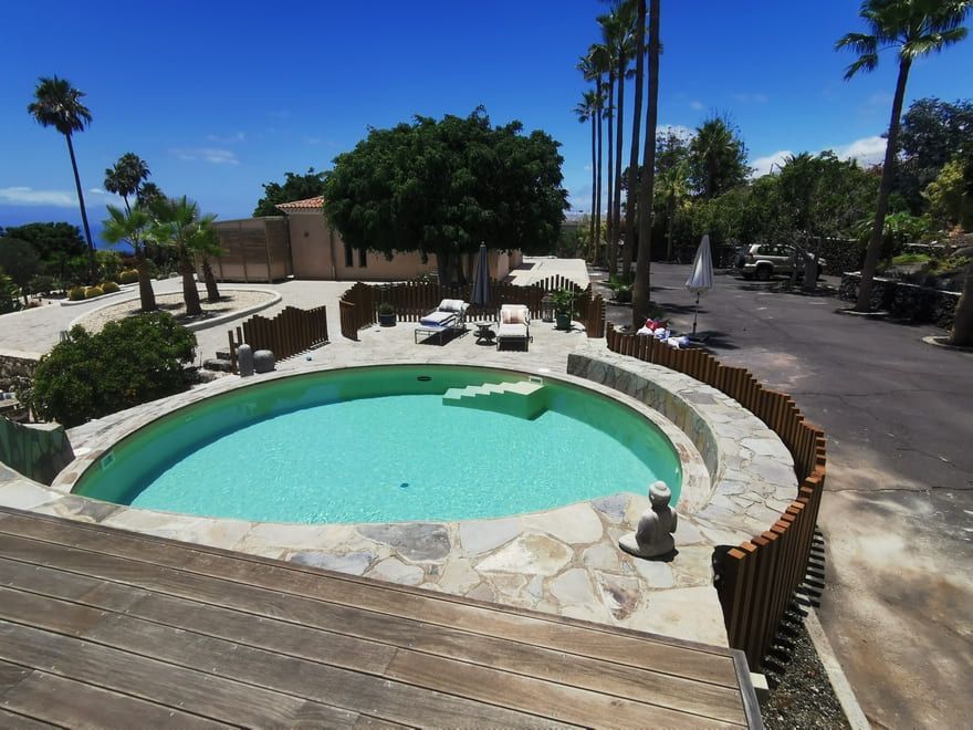 Patio del complejo con una piscina circular de color turquesa, palmeras y terraza de madera bajo un cielo soleado.