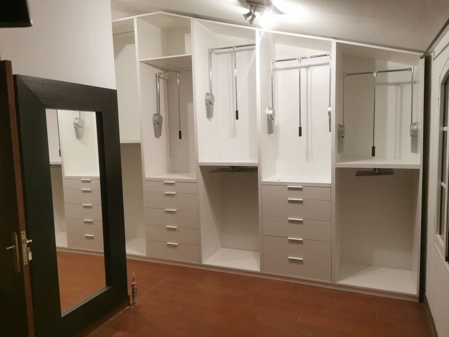 Vestidor con armarios blancos, cajones, barras para colgar ropa y un espejo de cuerpo entero sobre un suelo de madera.