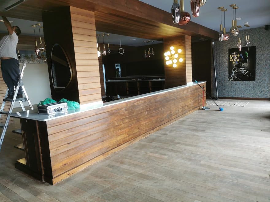 Interior de un bar vacío con mostrador revestido de madera, paredes negras, lámparas colgantes y un trabajador en una escalera a la izquierda.