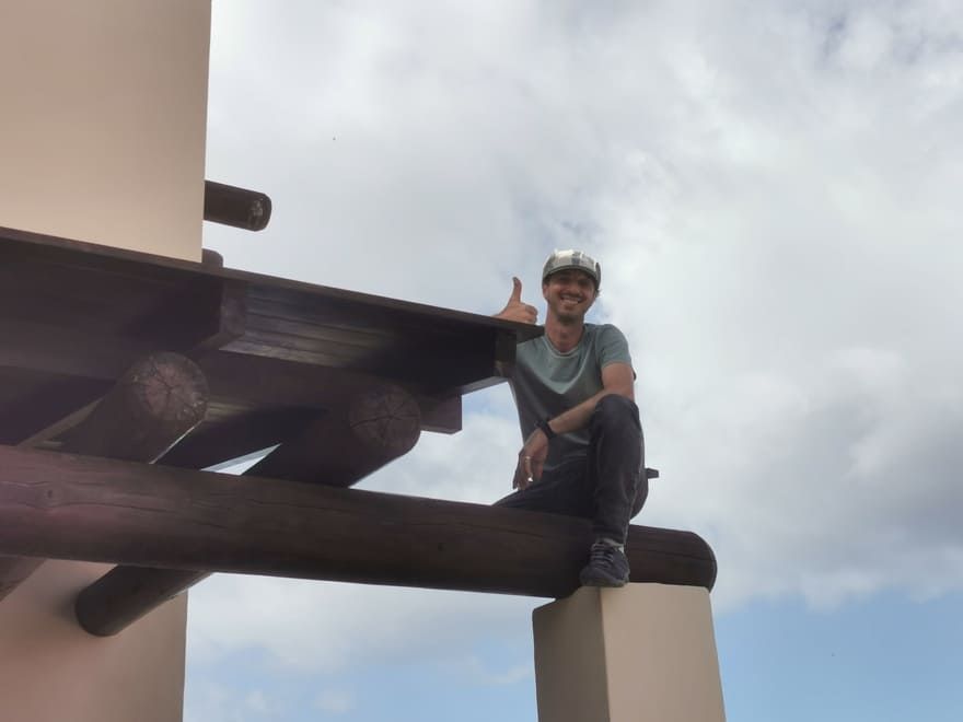 Una persona con casco está sentada sobre una gran viga en la azotea, sujetándose con una mano bajo un cielo nublado.