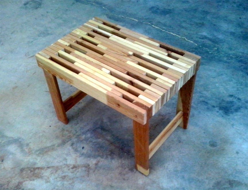 Taburete pequeño de madera con asiento de listones sobre un suelo de hormigón.