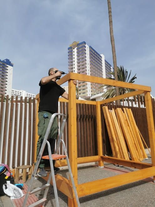 Una persona construye una estructura de madera en una terraza en la azotea, con una escalera y edificios de la ciudad al fondo.
