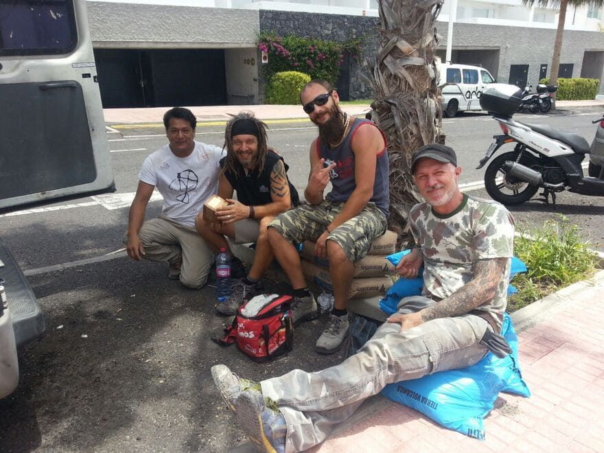 Cuatro personas sentadas en una acera bajo una palmera, sonriendo junto a una moto y una bolsa aparcadas.