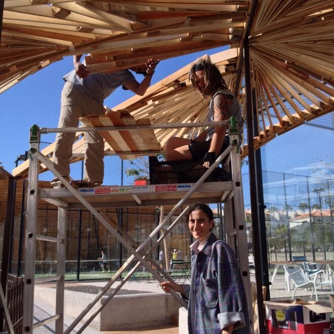 Trabajadores construyen un toldo de madera sobre andamios en un soleado patio al aire libre.