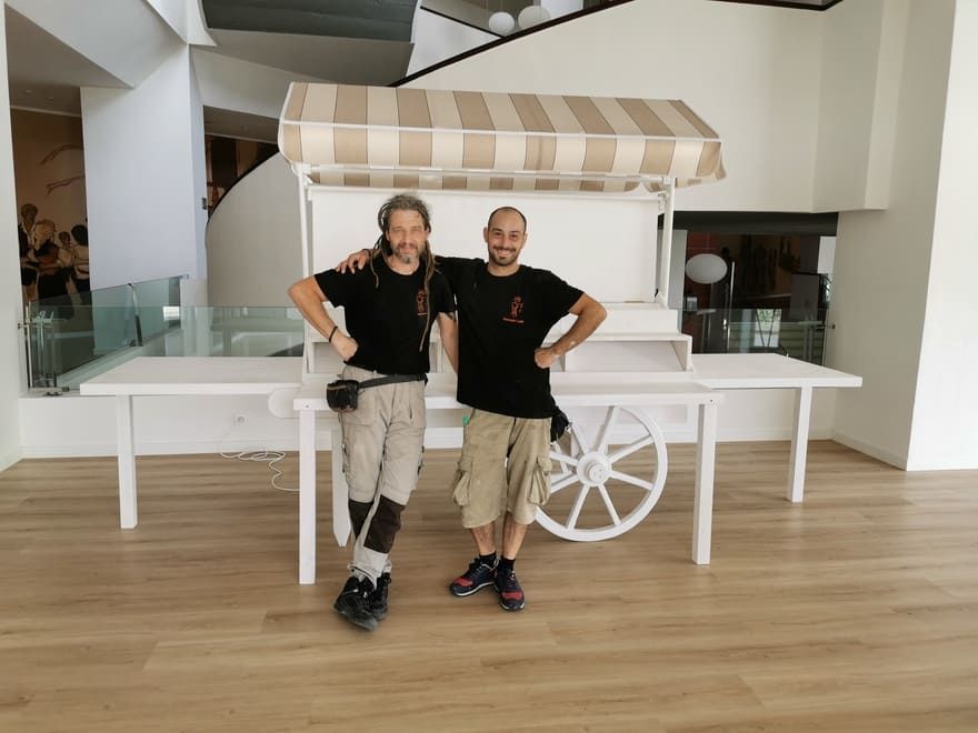 Dos personas posando delante de un carrito blanco con un toldo a rayas en un espacio interior luminoso.