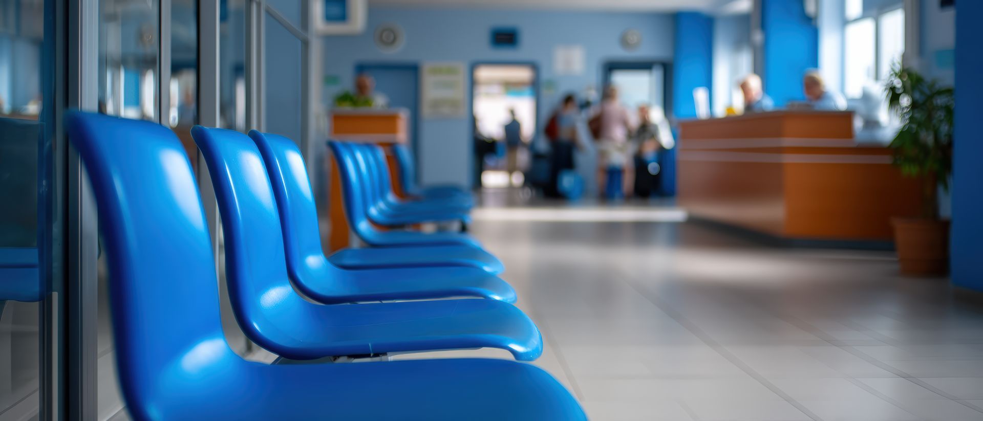 Des chaises bleues dans une salle d'attente d'hôpital, avec des silhouettes floues près d'un comptoir d'accueil.