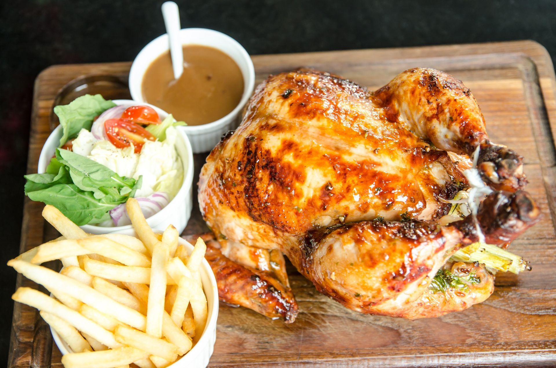 Pollo asado con salsa, ensalada y patatas fritas sobre una tabla de madera.