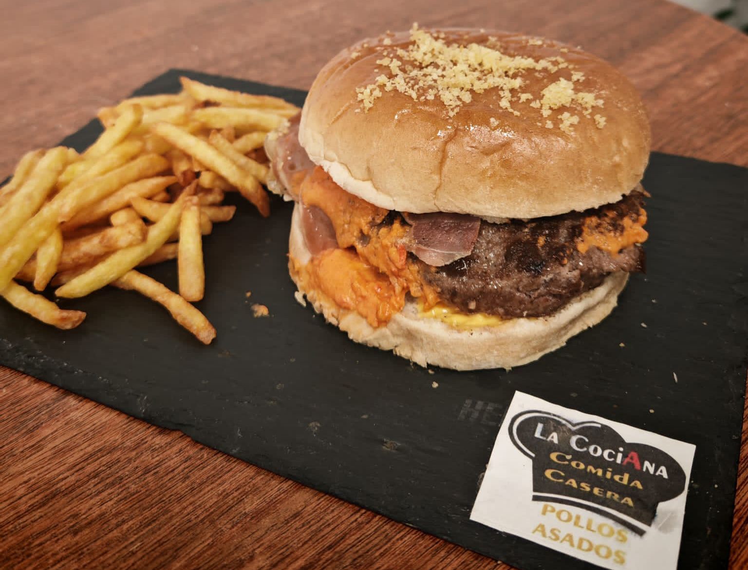 Hamburguesa con papas fritas en una pizarra negra. Incluye una hamburguesa, salsa de naranja y pan. Las papas están doradas. Hay una servilleta cerca.