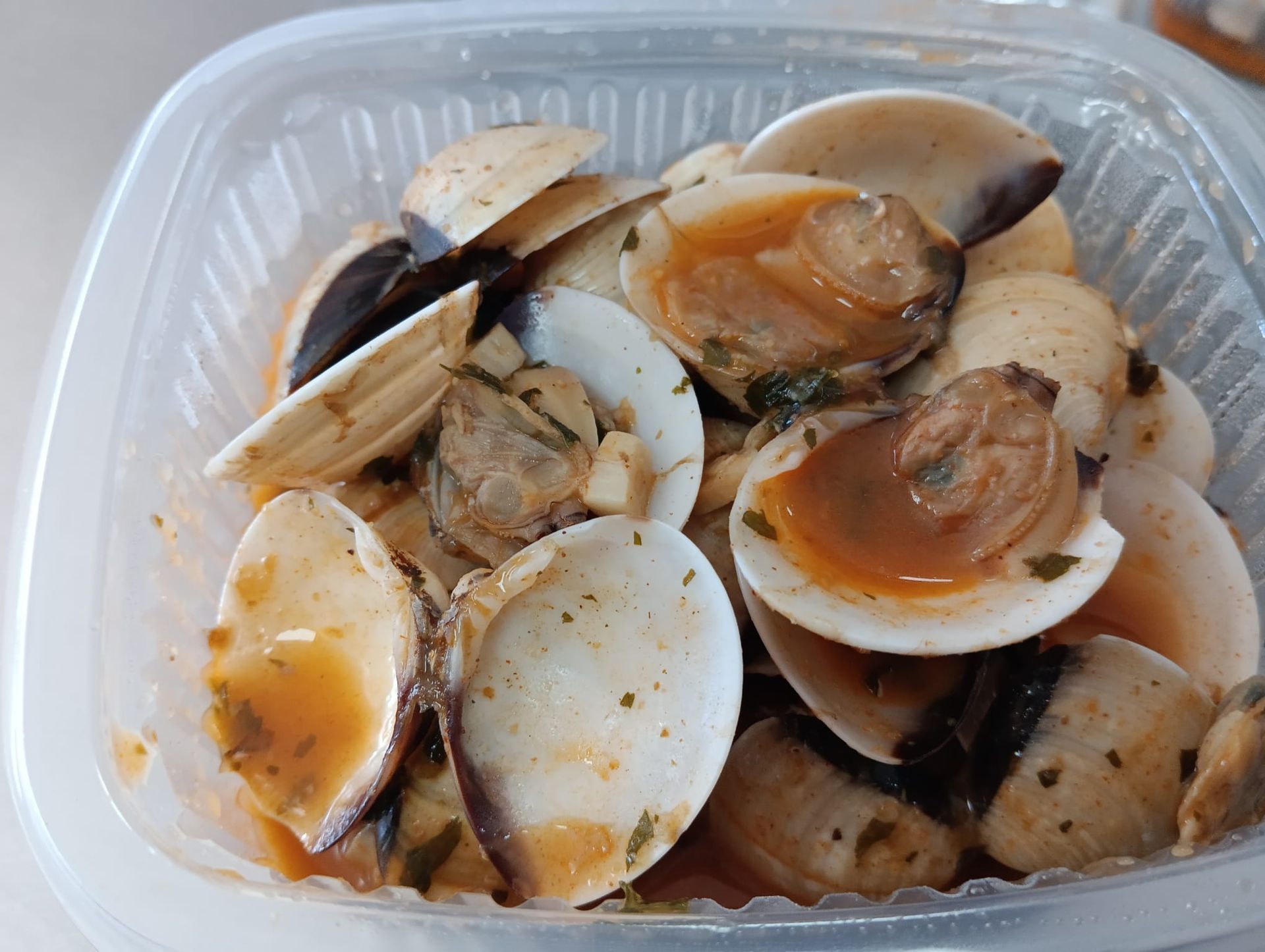 Almejas en un recipiente de plástico transparente, con salsa, hierbas y conchas abiertas.
