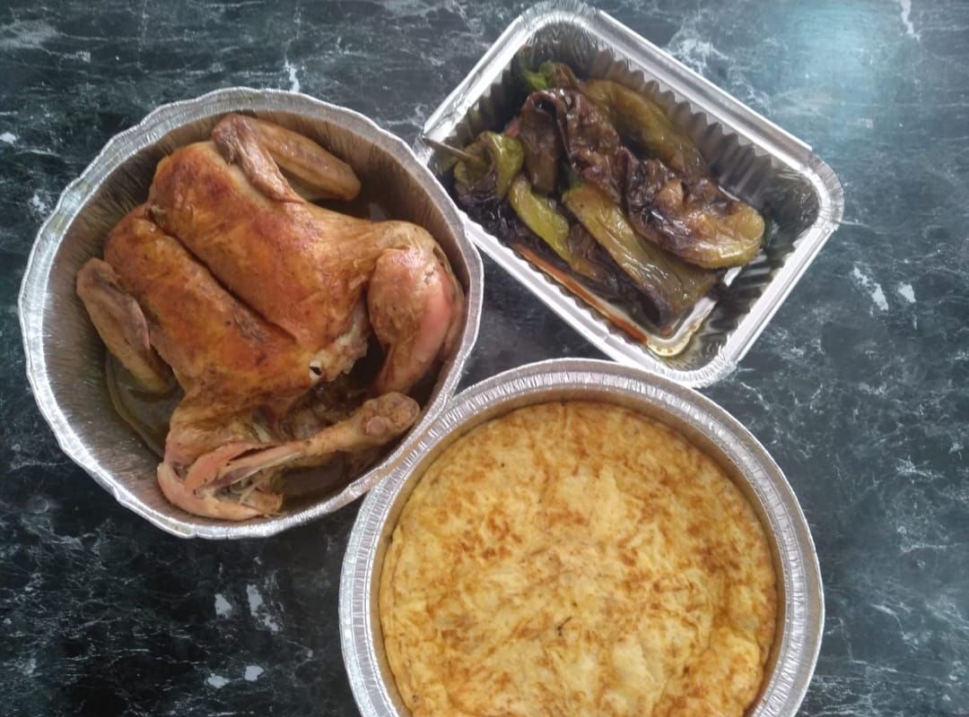 Pollo asado, berenjena y quiche en recipientes de aluminio sobre una superficie oscura.