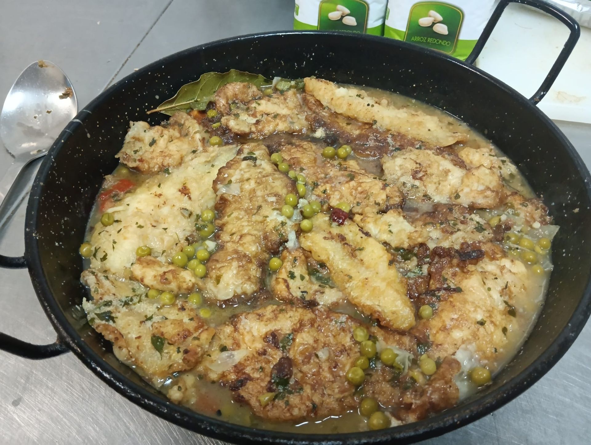 Guiso de pescado en sartén negra con guisantes, hierbas y una hoja de laurel.