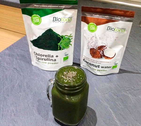 Un frasco de batido verde junto a dos bolsas de clorella y agua de coco.