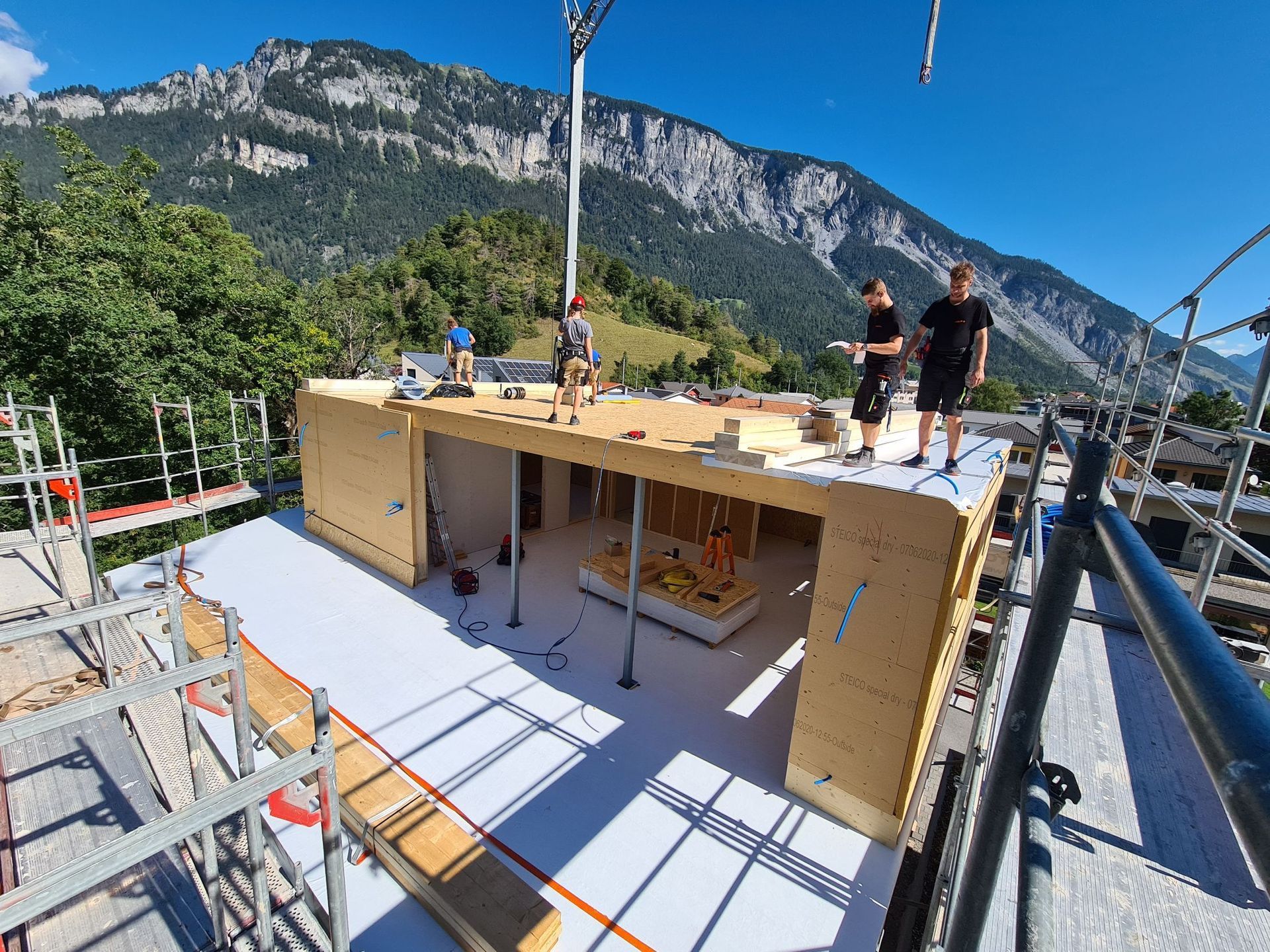 Zwei Männer stehen auf einem im Bau befindlichen Gebäude. Im Hintergrund sind Berge zu sehen.