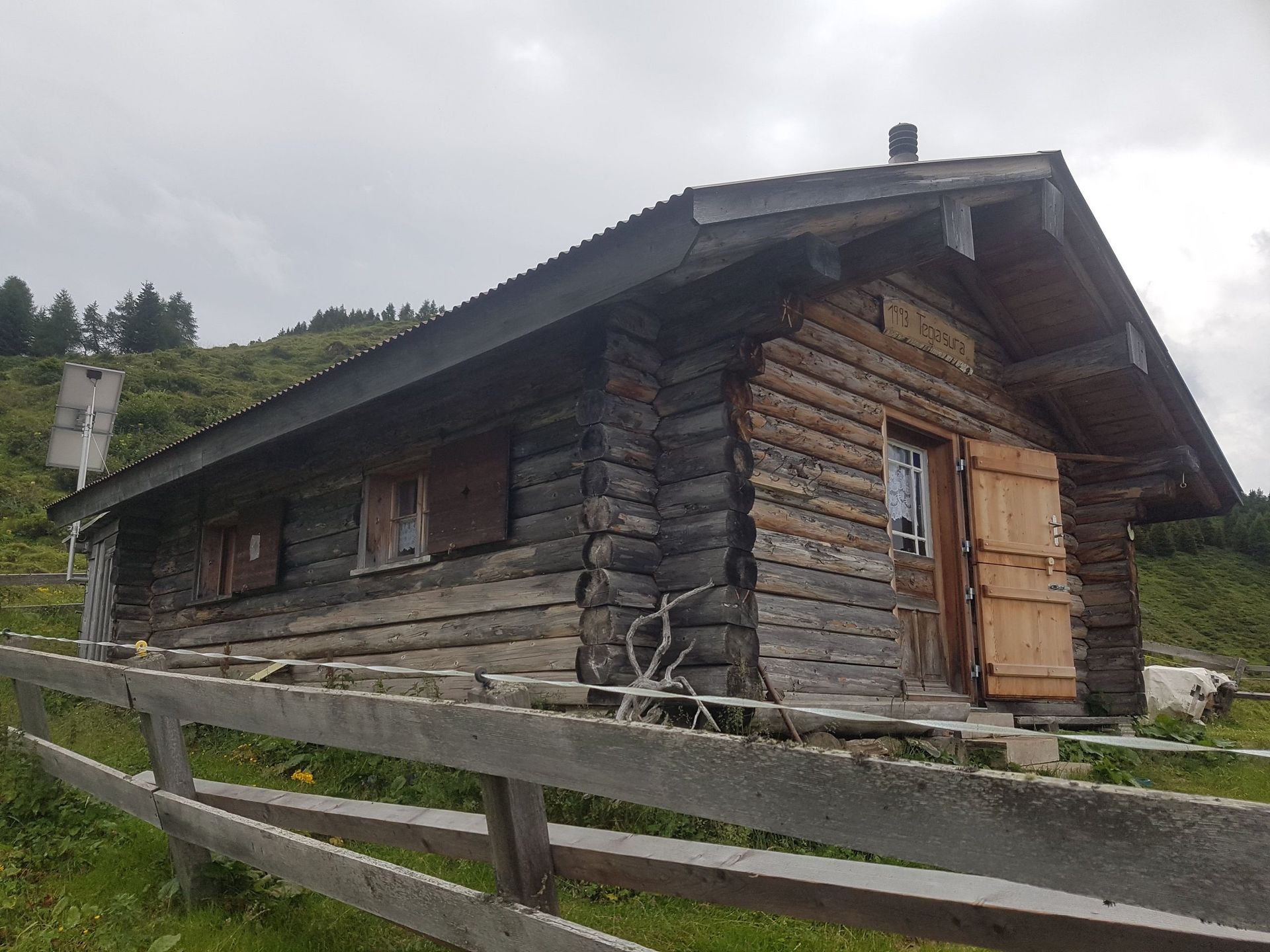 Auf einem grasbewachsenen Hügel befindet sich hinter einem Holzzaun eine kleine Blockhütte.
