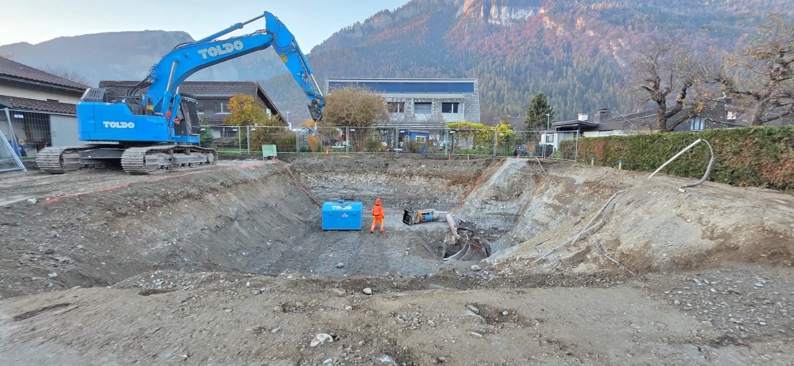 Baustelle mit einem blauen Bagger, Erdarbeiten und Bergen im Hintergrund.