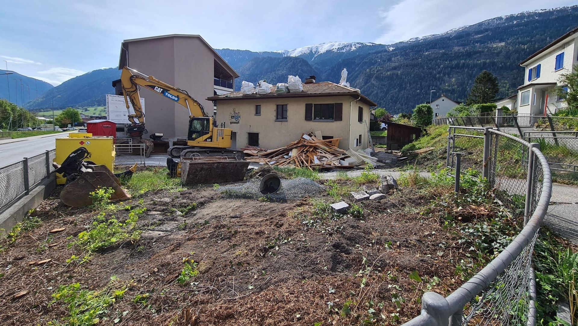 Ein Haus wird abgerissen, im Vordergrund ist ein gelber Bagger zu sehen.