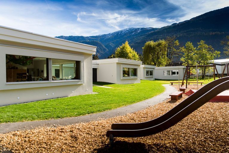 Eine Rutsche auf einem Spielplatz vor einem Gebäude mit Bergen im Hintergrund.