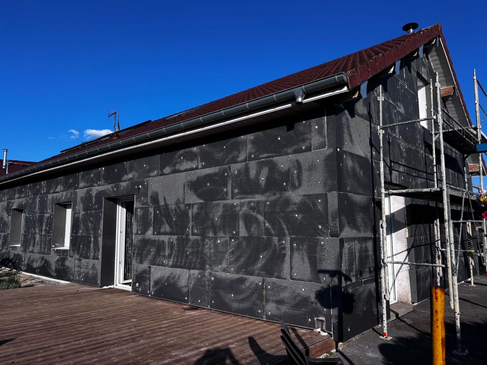 Façade d'une maison en cours d'isolation avec des échafaudages et un toit de tuiles rouges sous un ciel bleu clair