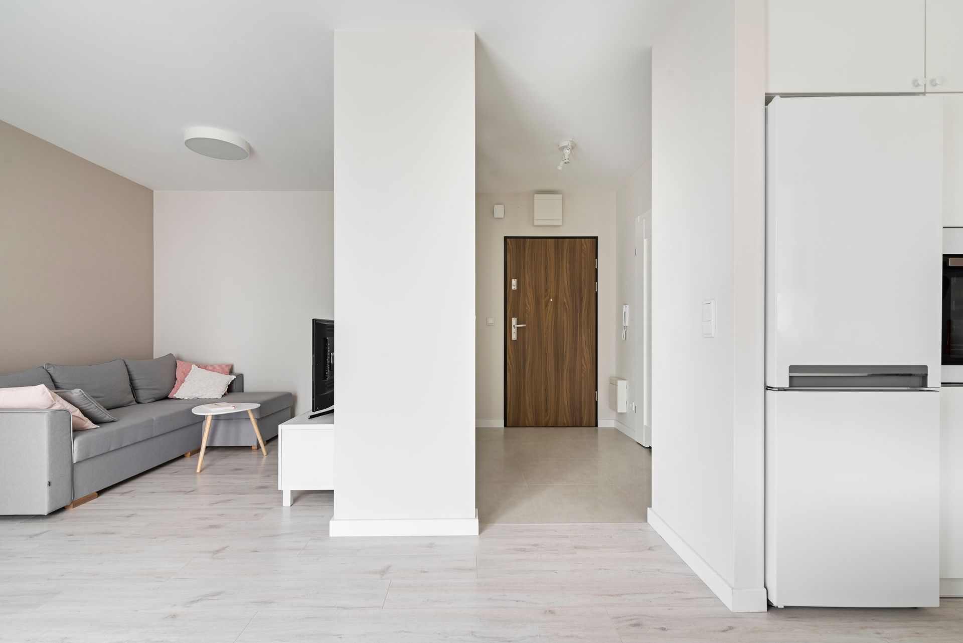 Salon minimaliste blanc avec canapé gris, porte d'entrée en bois et réfrigérateur