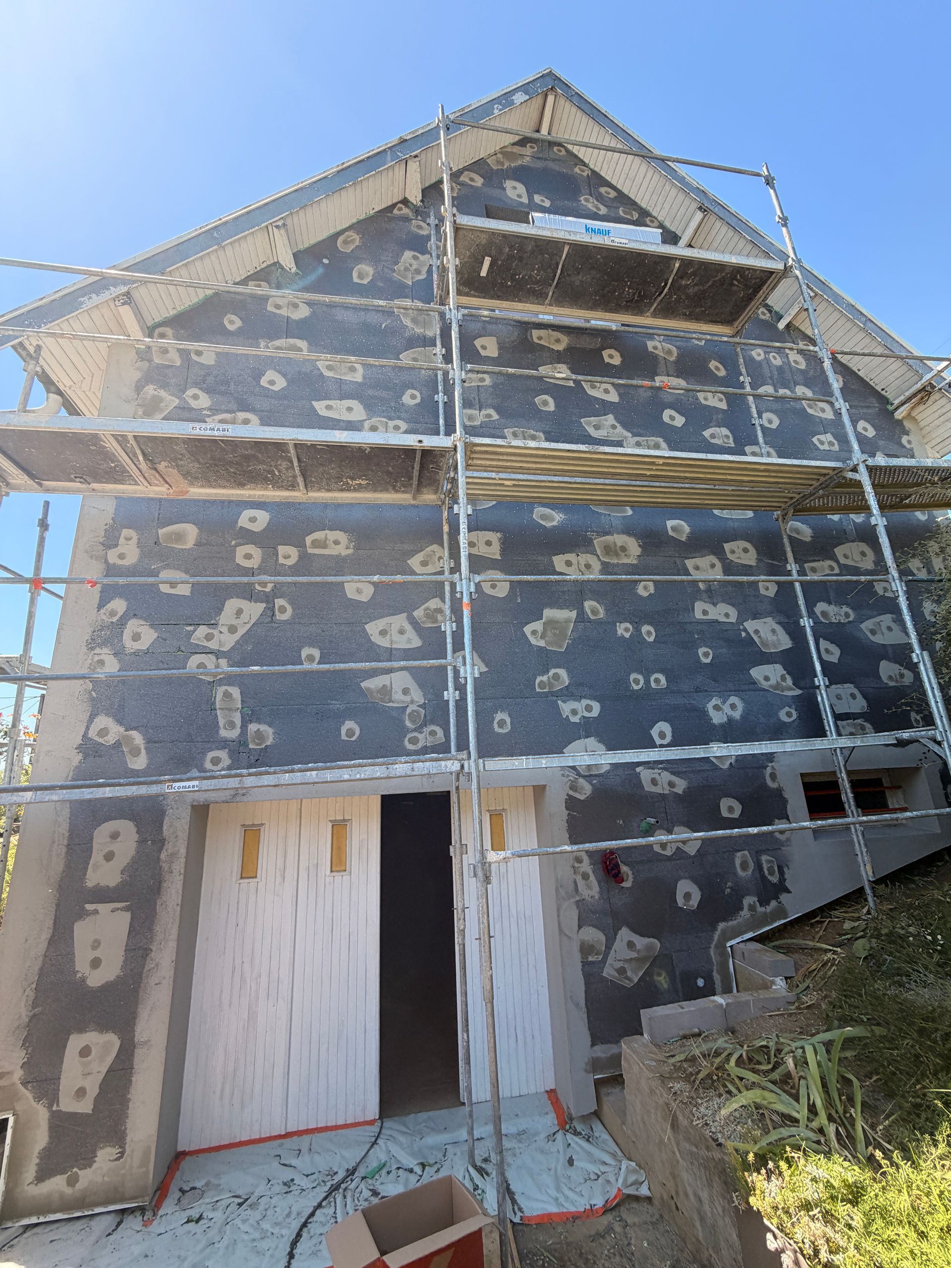 Maison grise en rénovation avec échafaudages, structures métalliques et ciel bleu clair