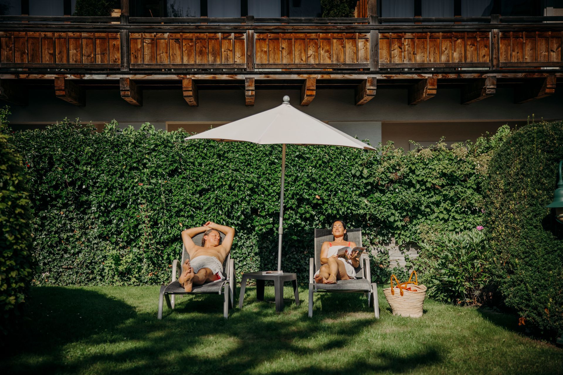 Zwei Personen sitzen in Liegestühlen unter einem Sonnenschirm in einem Garten.