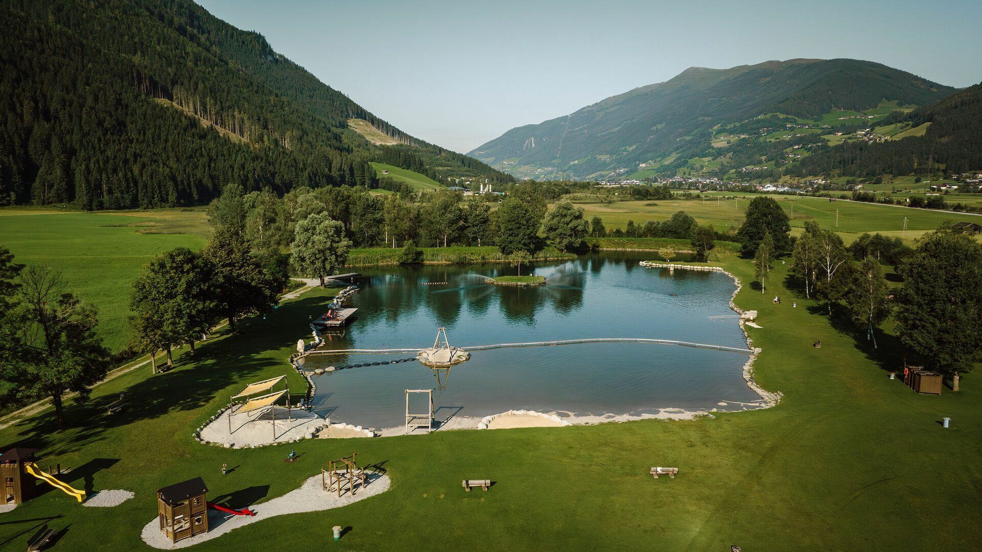 Eine Luftaufnahme eines von Bäumen und Gras umgebenen Sees mit Bergen im Hintergrund.
