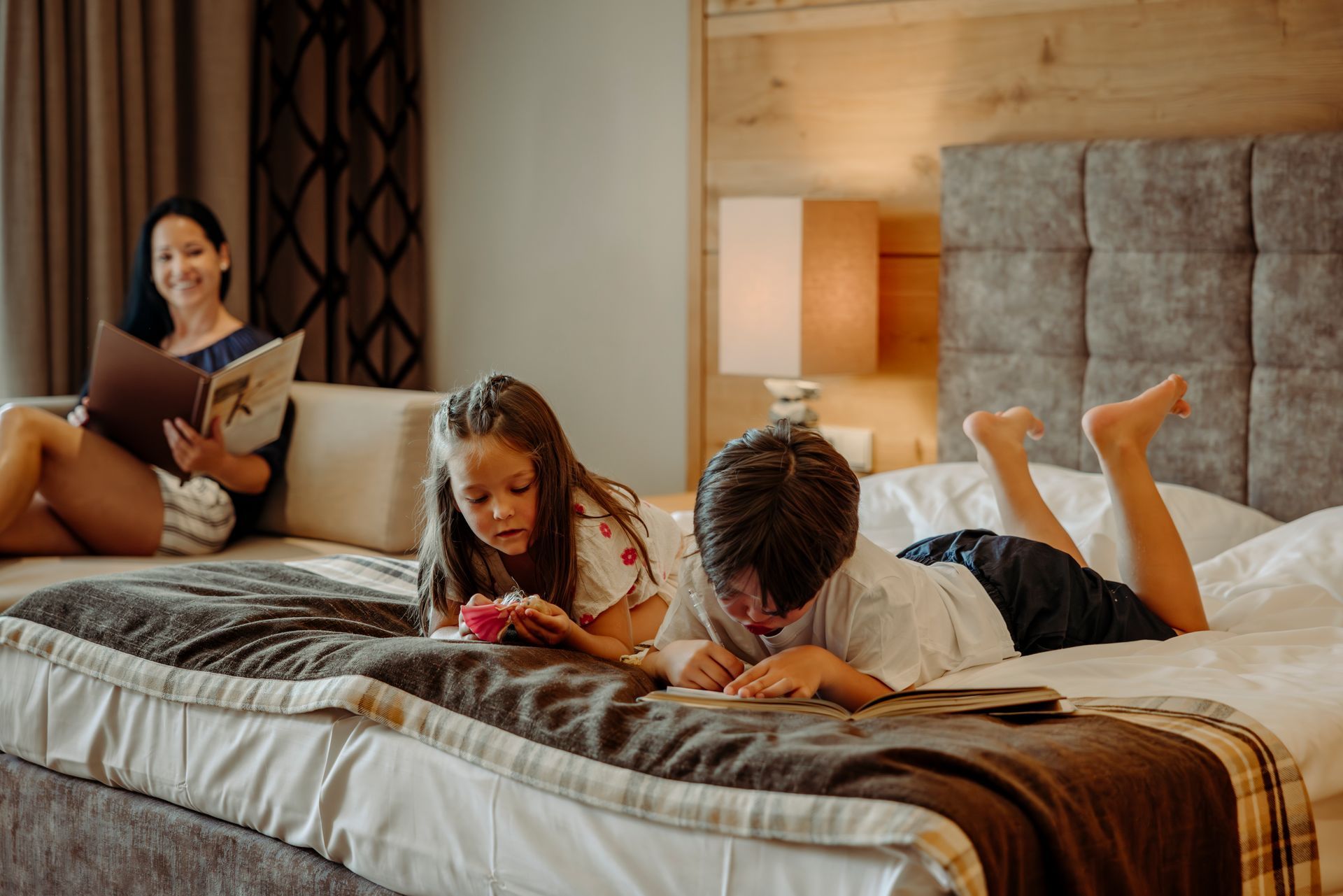 Eine Frau sitzt auf einer Couch und liest ein Buch, während zwei Kinder auf einem Bett liegen.
