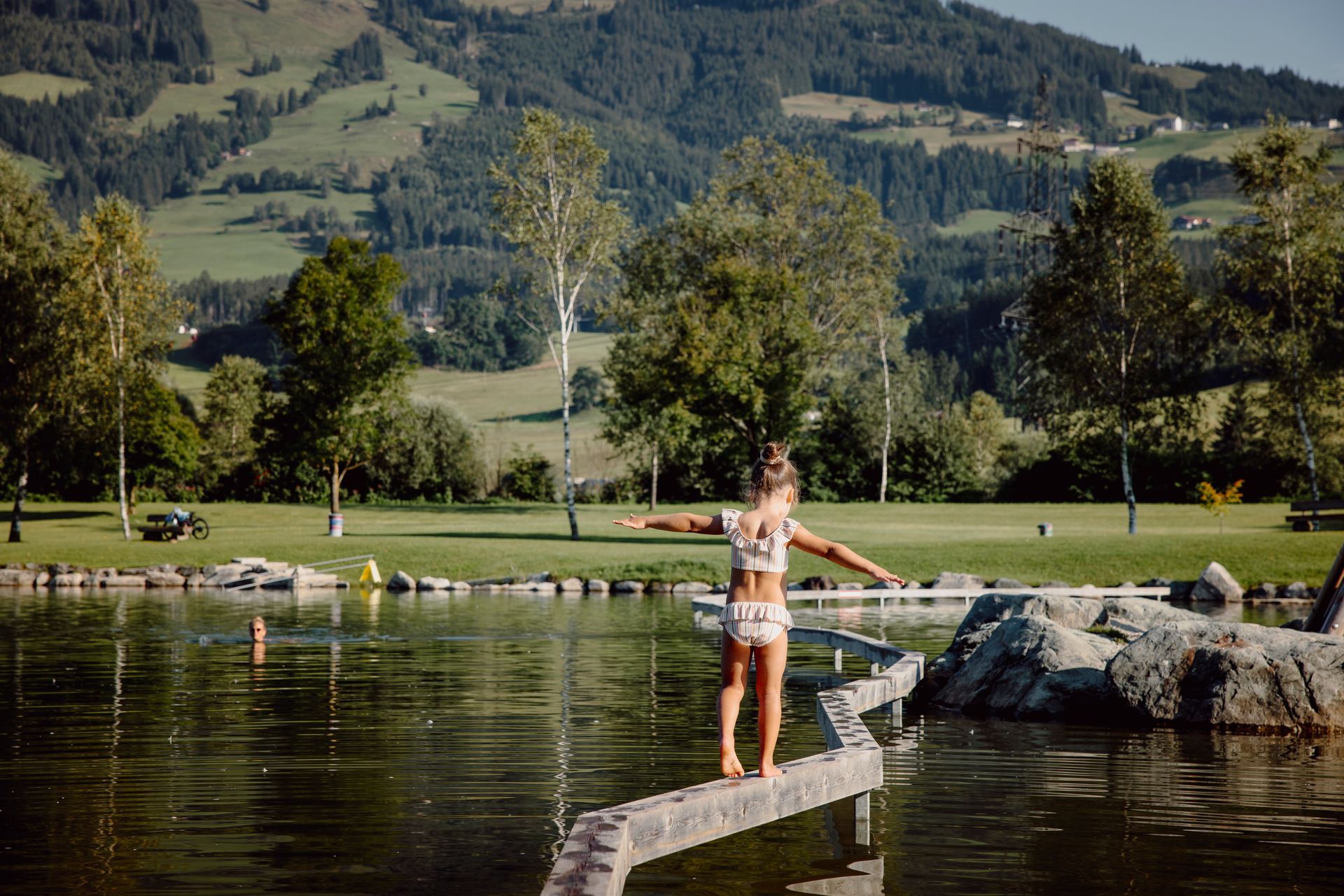 Ein Mädchen balanciert auf einem Holzsteg über den Naturbadesee in Hollersbach im Salzburger Land.