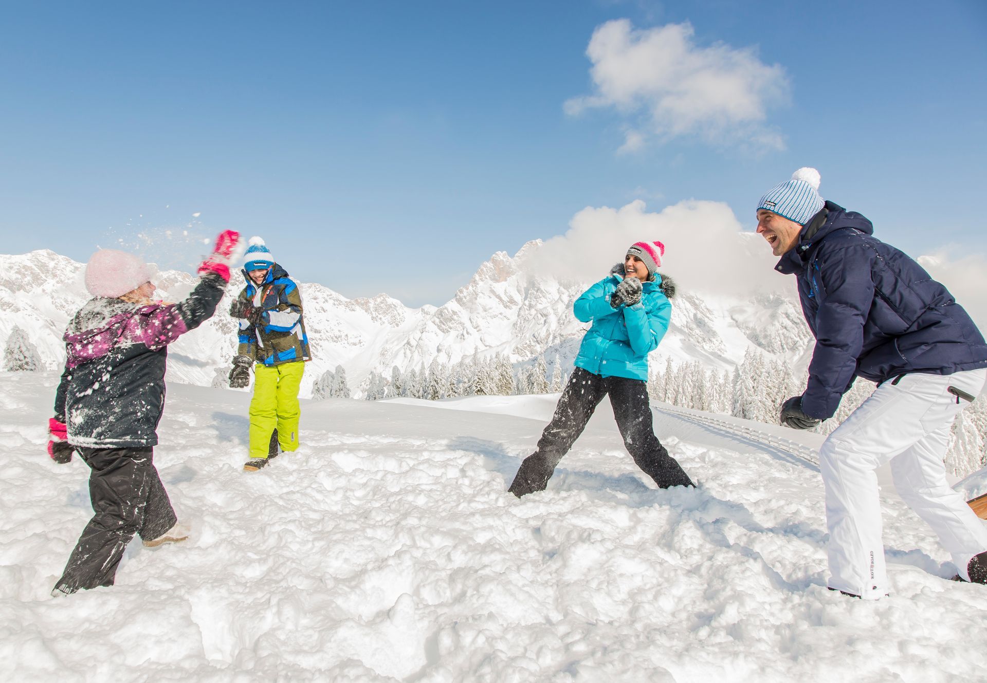 Vier Menschen in Winterkleidung spielen auf der Maria Alm im tiefen Schnee, im Hintergrund sind Berge zu sehen