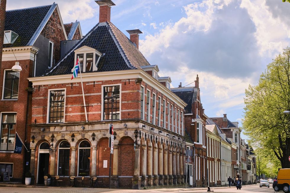 Rood bakstenen gebouw met pilaren en een donker dak, met een vlag in de lucht, in een zonnige straat in een Europese stad.