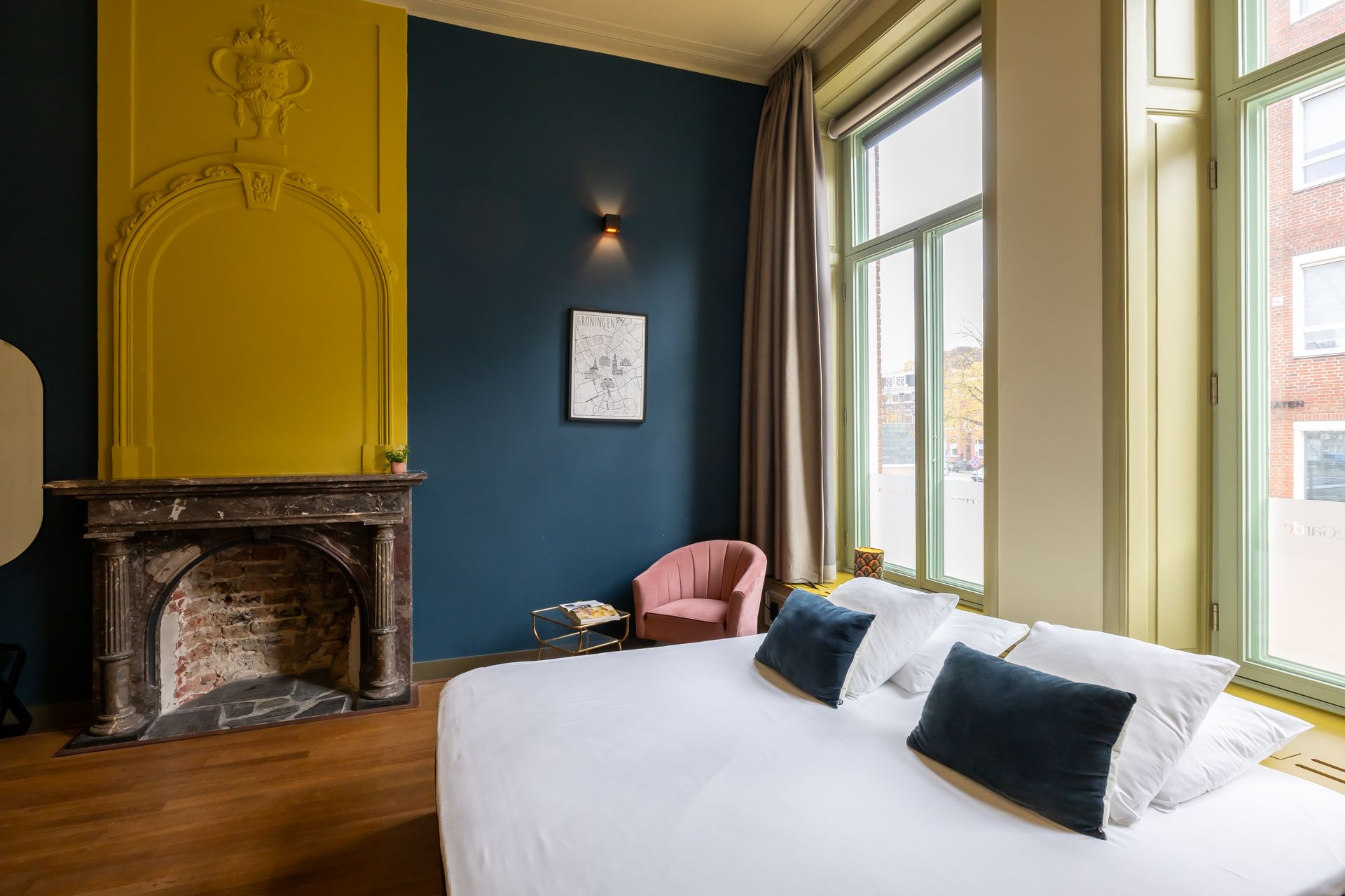 Slaapkamer met een blauwe muur, gele accenten, open haard, grote ramen en een bed met blauwe kussens.