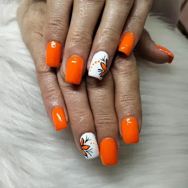 Uñas cuadradas de color naranja con detalles en blanco que presentan diseños florales sobre una superficie blanca esponjosa.