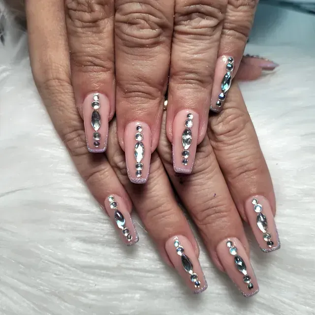 Manos con uñas largas y cuadradas de color rosa claro. Cada uña tiene una línea vertical de pedrería plateada.