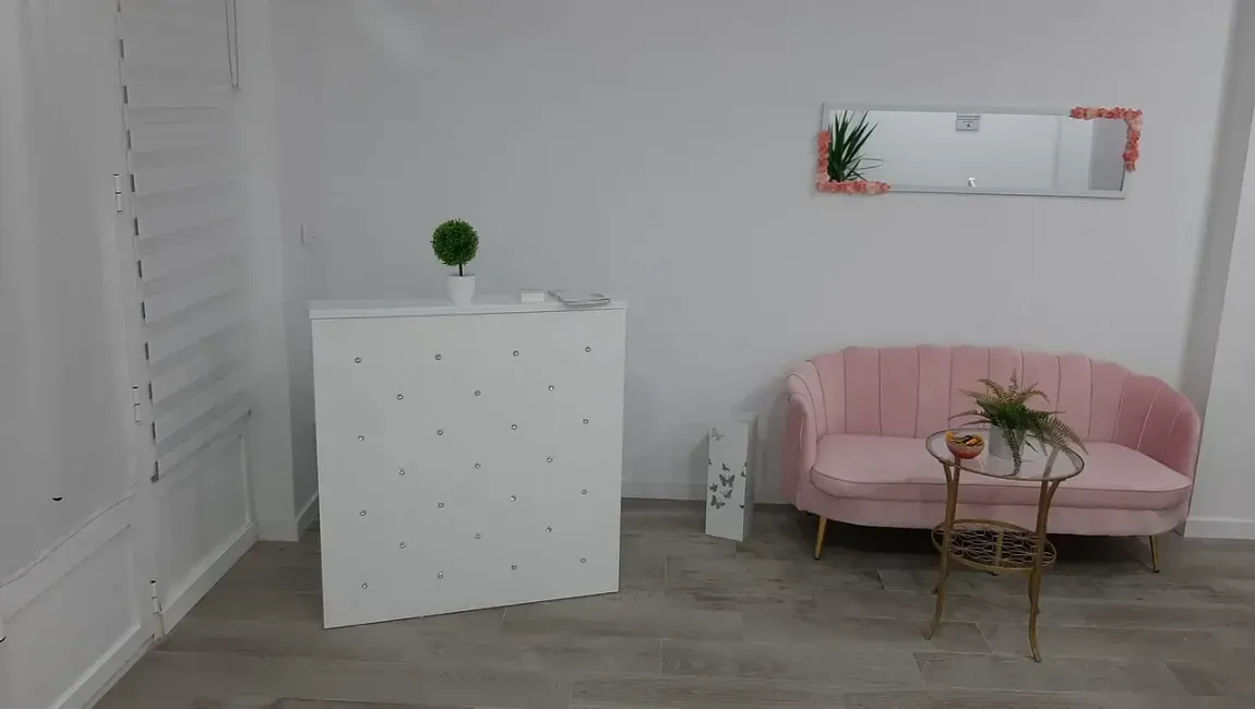 Habitación blanca con sofá rosa, mesa pequeña, escritorio blanco y espejo con arte.