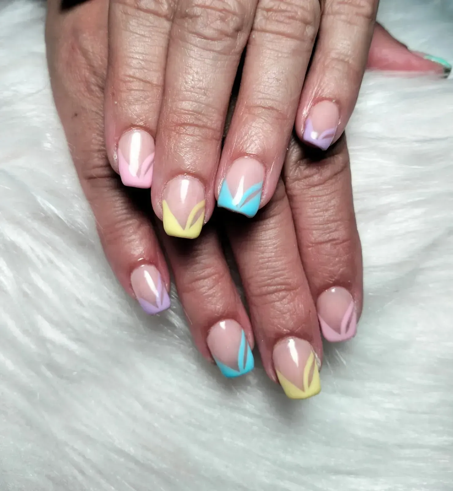 Uñas con punta francesa en colores pastel con diseños florales sobre una superficie blanca y esponjosa.