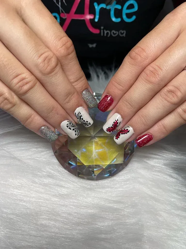 Manos de mujer con uñas cuidadas. Las uñas combinan brillo, blanco con detalles negros y rojo. Se encuentran sobre un soporte decorativo de cristal.